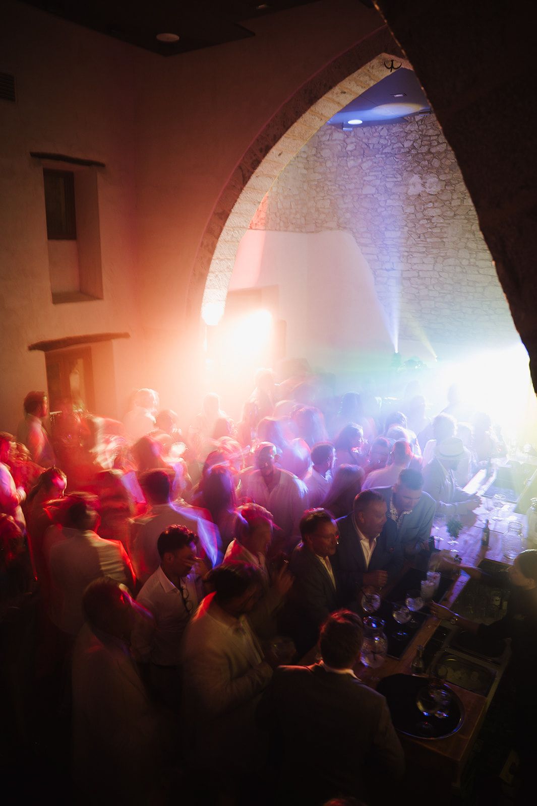 Wedding guests dancing under stone archway with colorful DJ lights at lively indoor after-party.