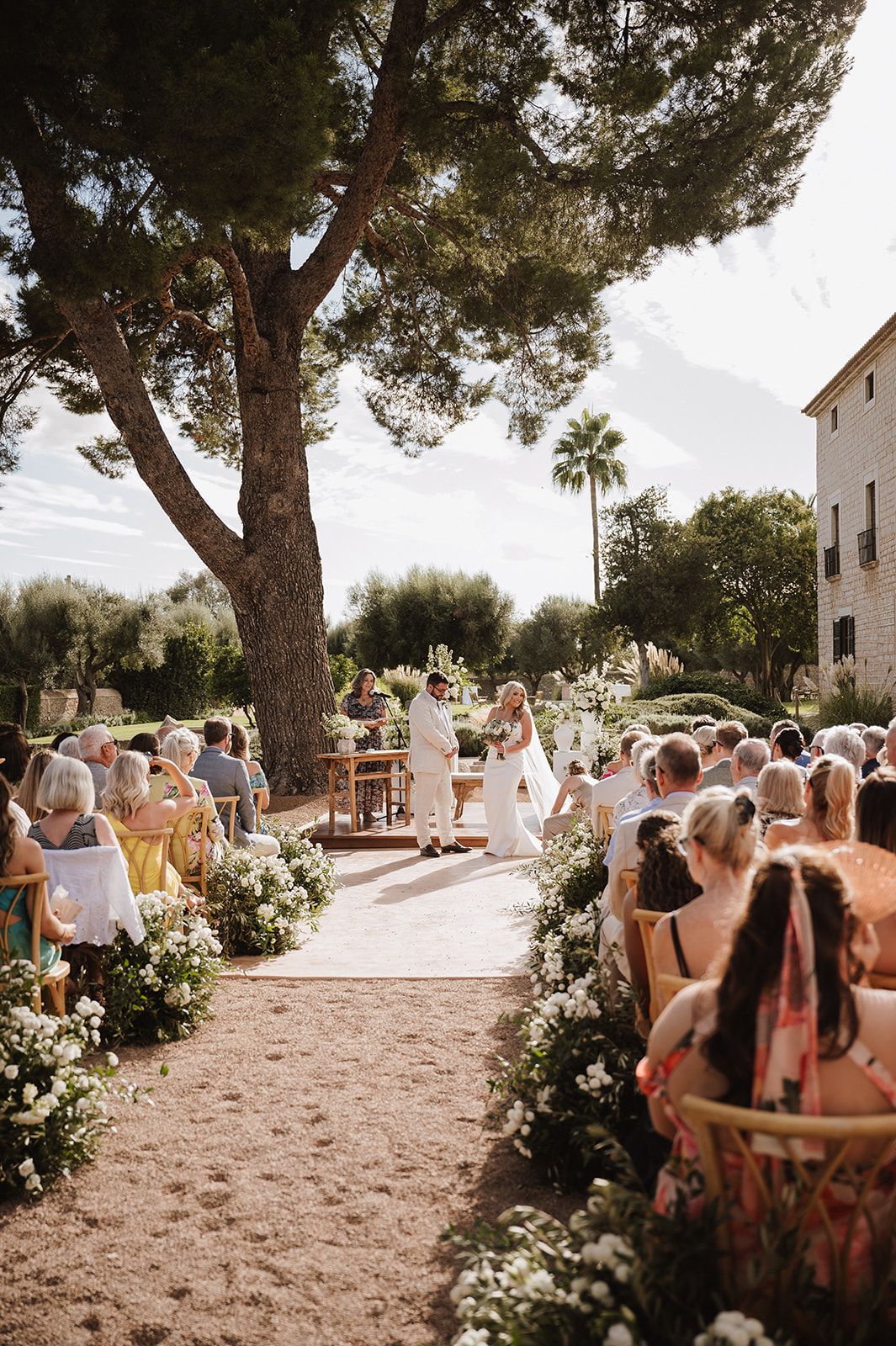 Guests seated at outdoor wedding ceremony beneath pine trees at Mediterranean venue.