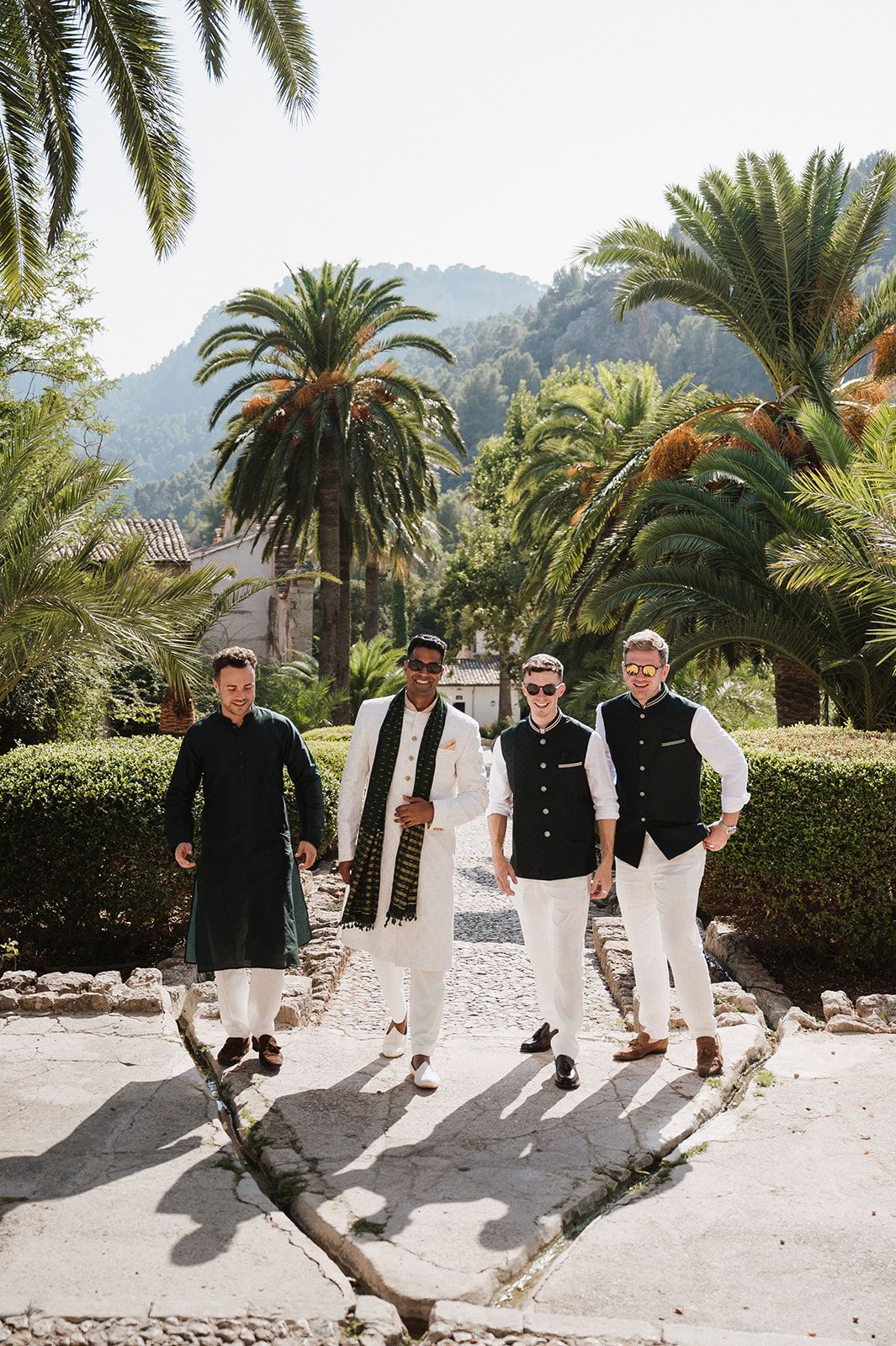 Four men in Indian wedding outfits walking together on stone path surrounded by tropical palm trees.