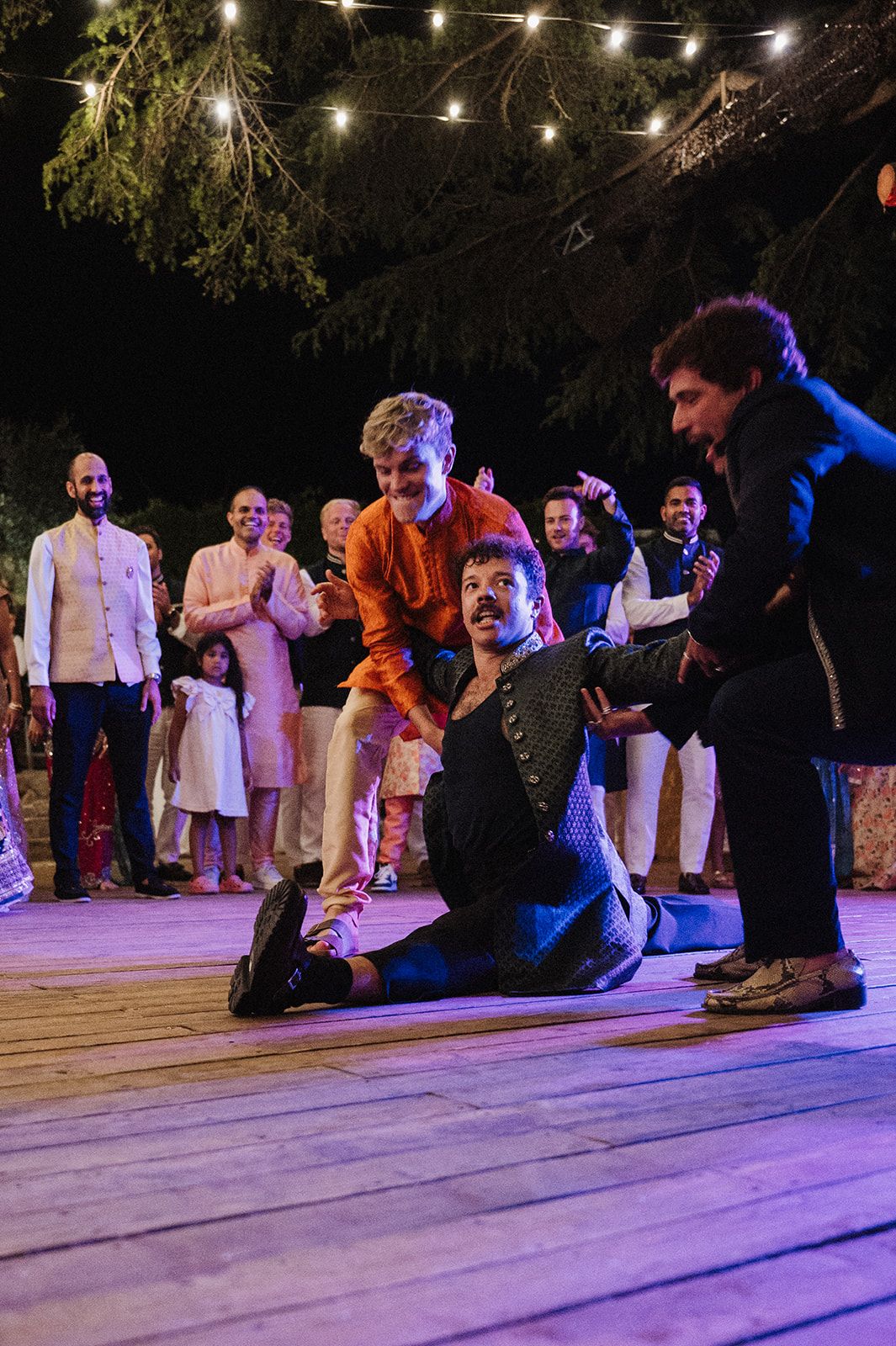 Energetic wedding guests dancing and celebrating on outdoor dance floor under string lights at night.