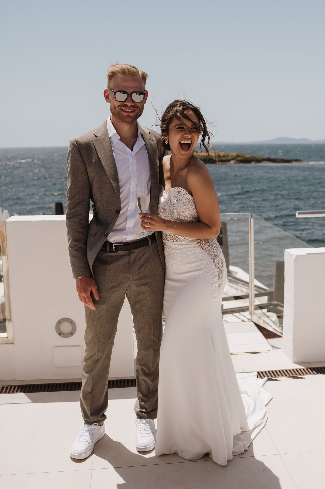 Smiling bride in white lace gown and groom in tan suit pose together on a sunny oceanfront terrace.