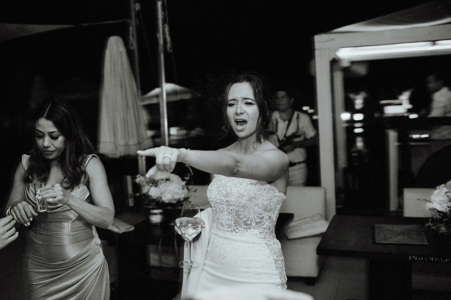 Bride in strapless lace gown dances energetically with bridesmaid at outdoor wedding reception, black and white.