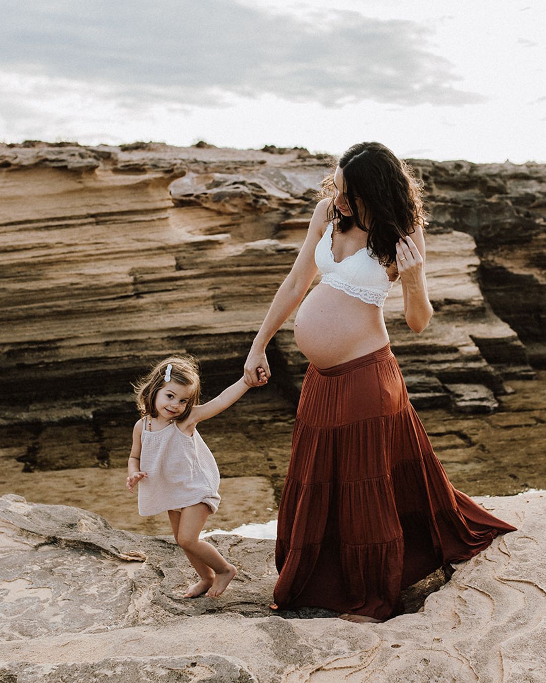 Pregnant woman in white bralette and rust maxi skirt holds toddler girl's hand on rocky coastal cliffs.
