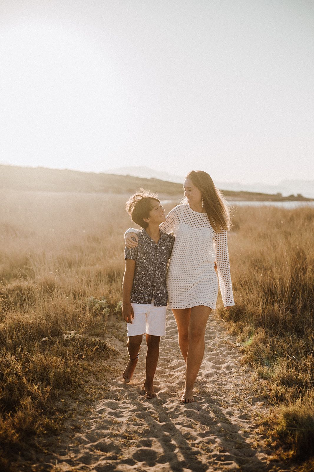 Mother and son walking barefoot on a sandy path through golden fields at sunset.