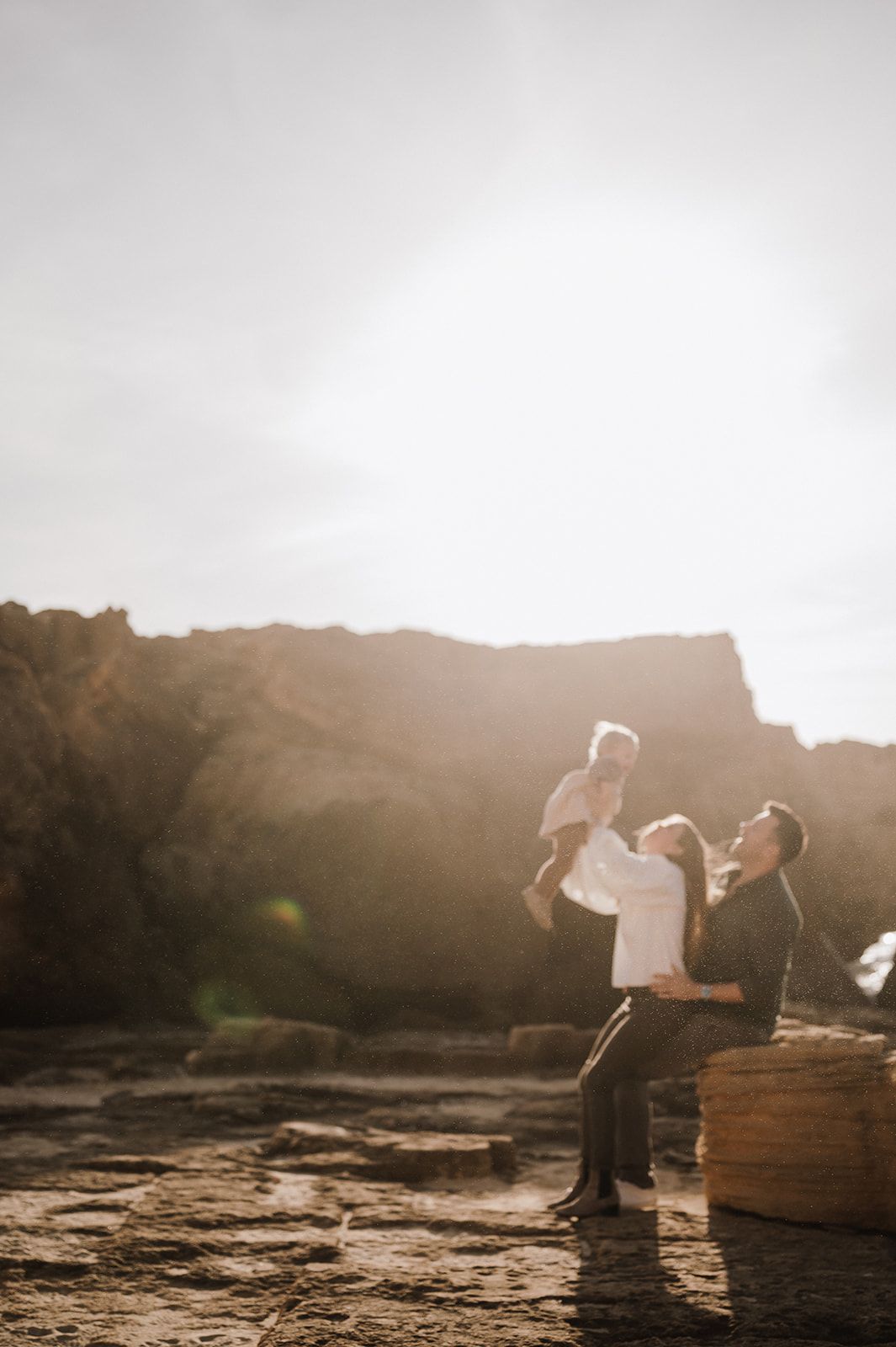 Family moment at golden hour on rocky coast, parent lifting baby while partner watches lovingly.