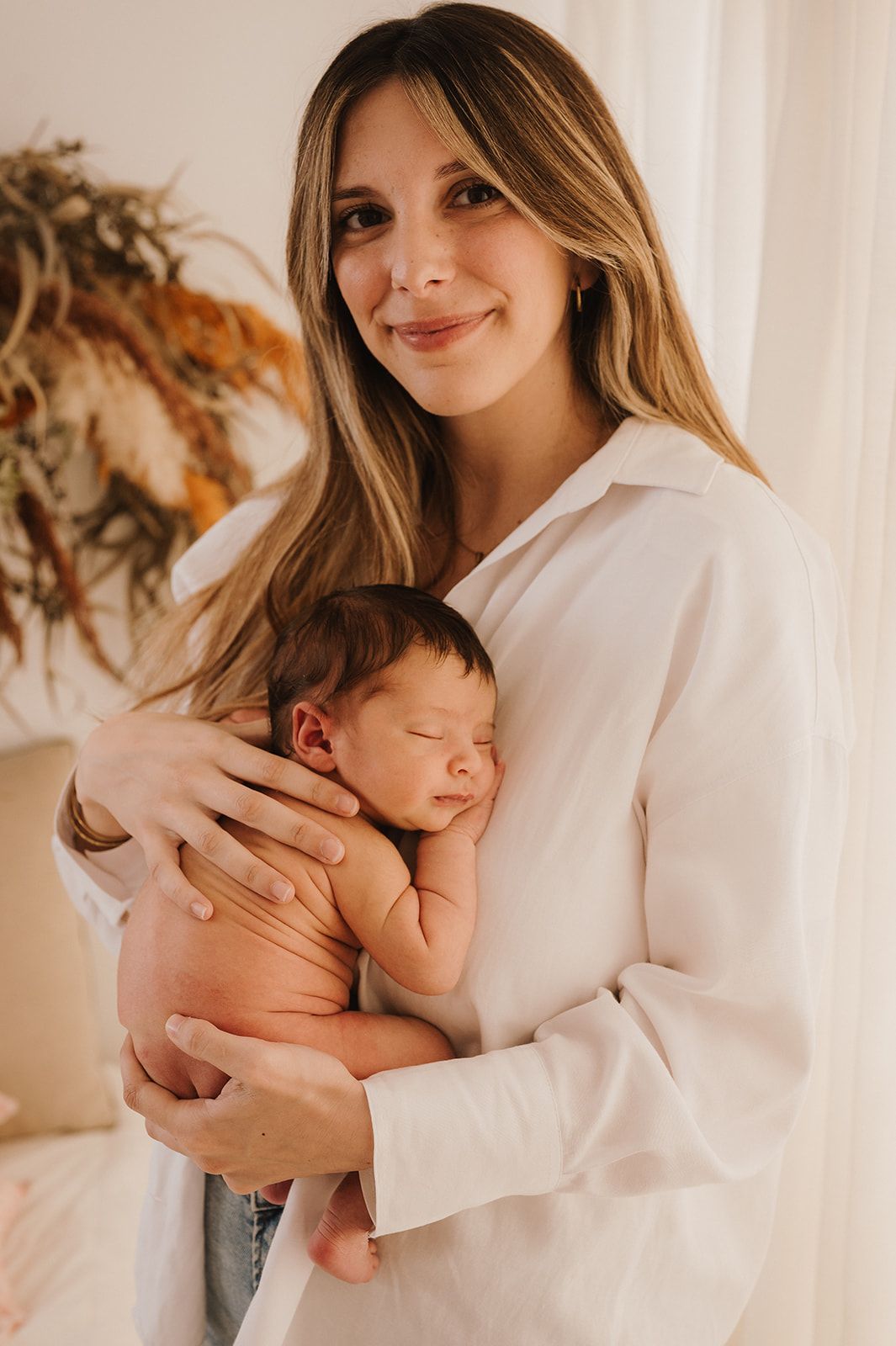 Smiling mother in white shirt gently cradling sleeping newborn baby against her chest indoors.