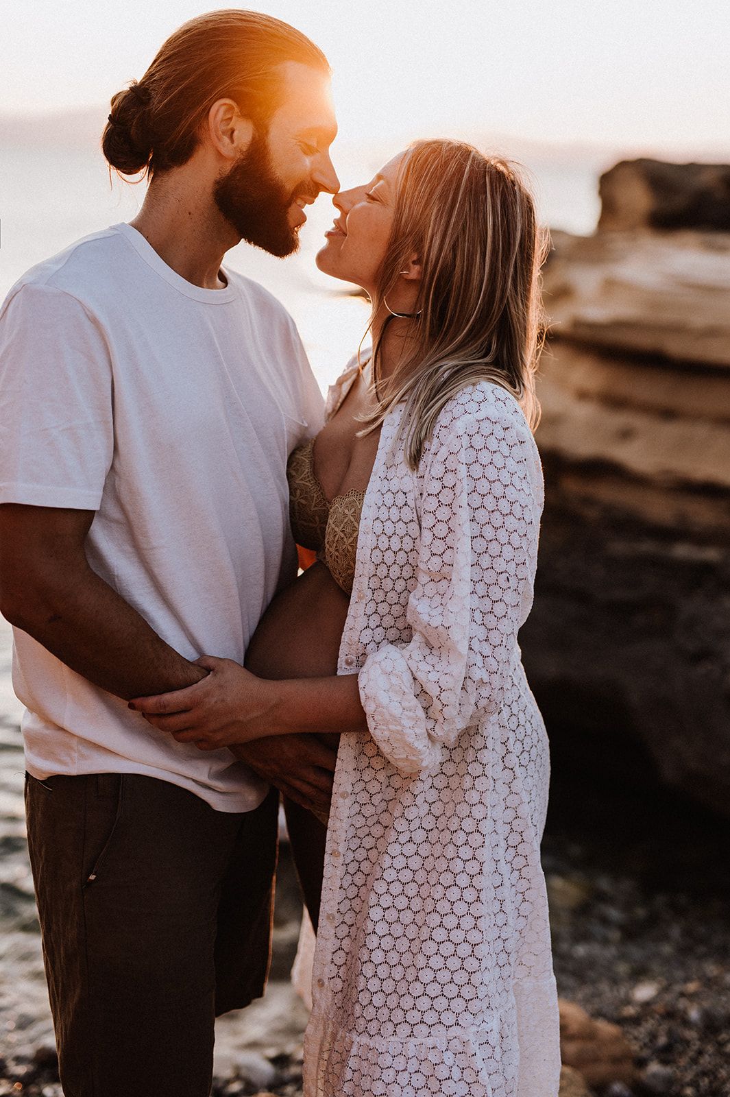 Couple sharing romantic moment at sunset by rocky beach, pregnant woman in white lace cardigan.