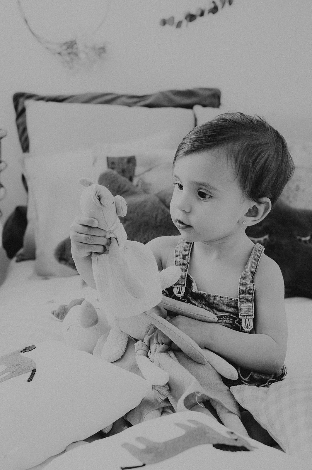 A curious toddler in denim overalls examines a stuffed animal toy while sitting on a bed, in black and white.