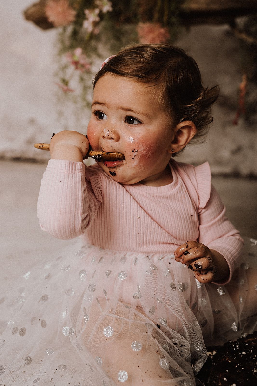 A baby girl in a pink onesie and tutu smiles with cake smeared on her face during a first birthday smash cake session.