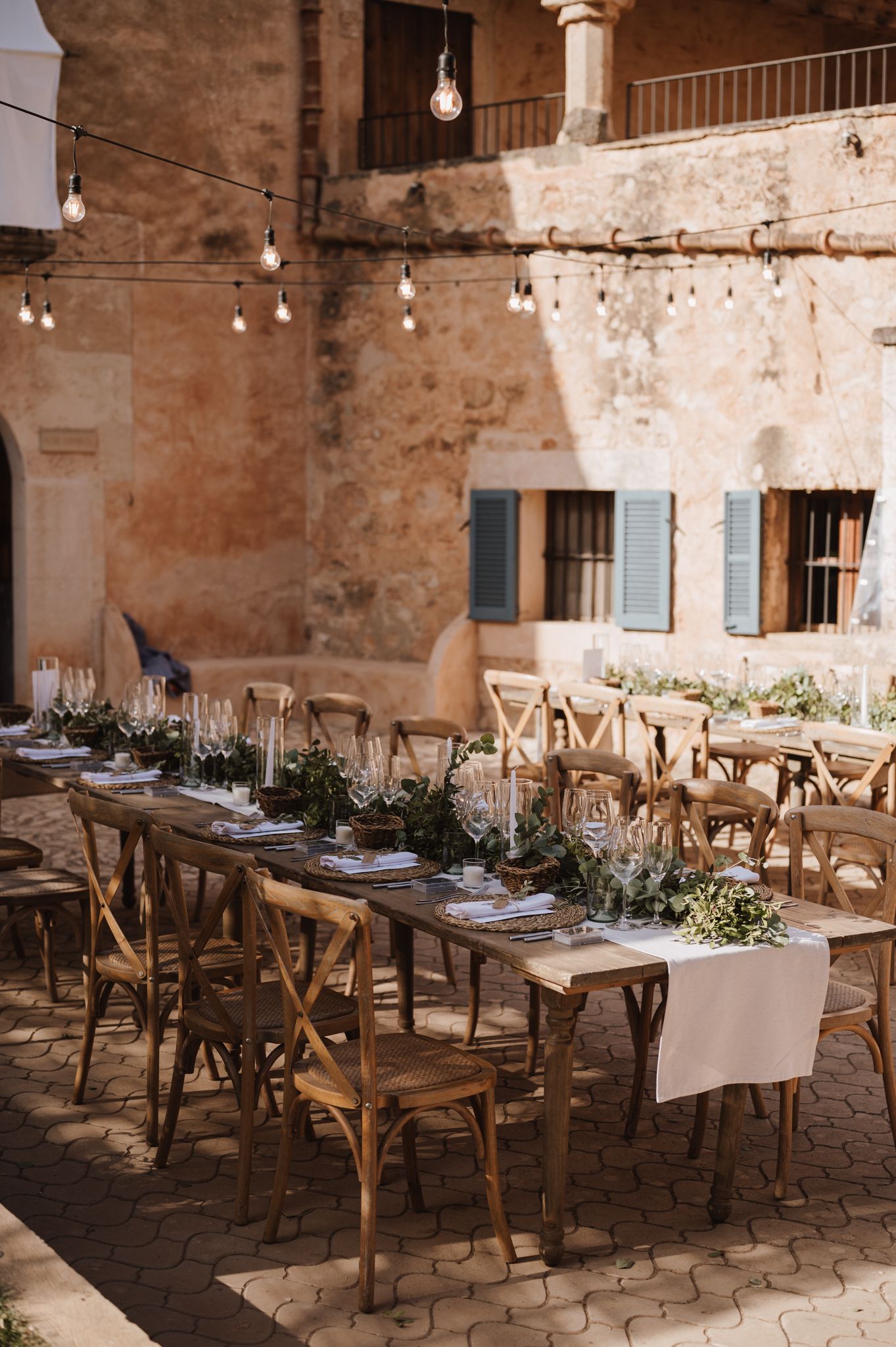 Rustic outdoor wedding reception table set with greenery runners and string lights at stone venue courtyard.
