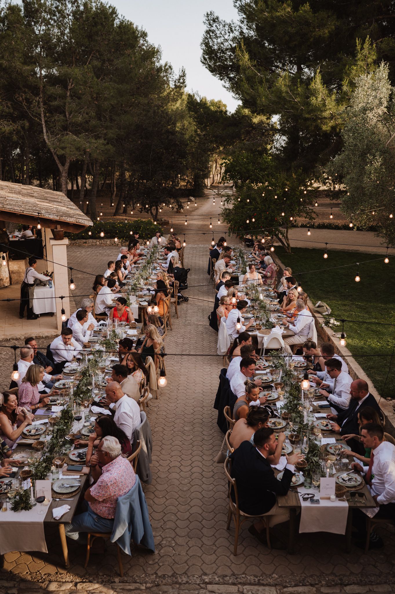 Aerial view of long outdoor wedding reception tables with guests dining under string lights surrounded by pine trees.