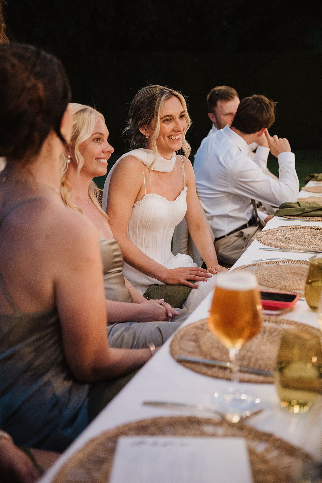 Radiant bride in strapless gown laughs with bridesmaids in sage dresses during outdoor wedding reception dinner.