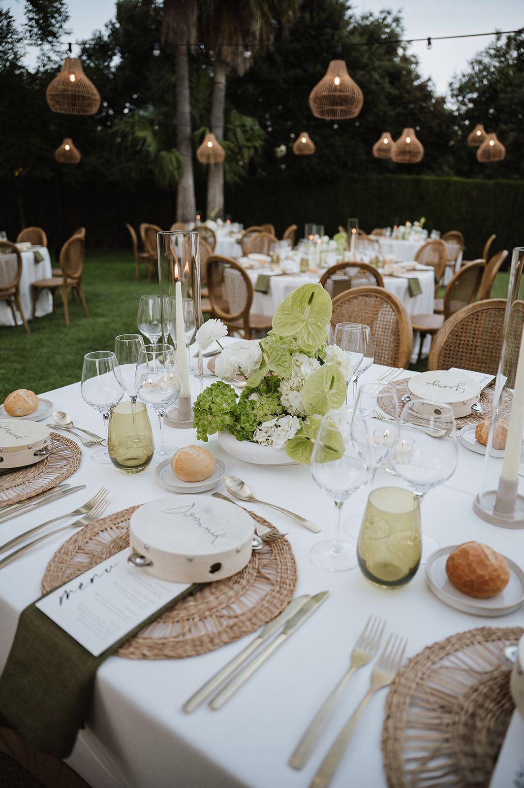Elegant outdoor wedding reception tables with wicker chargers, green glassware, white floral centerpieces, and rattan pendant lights.