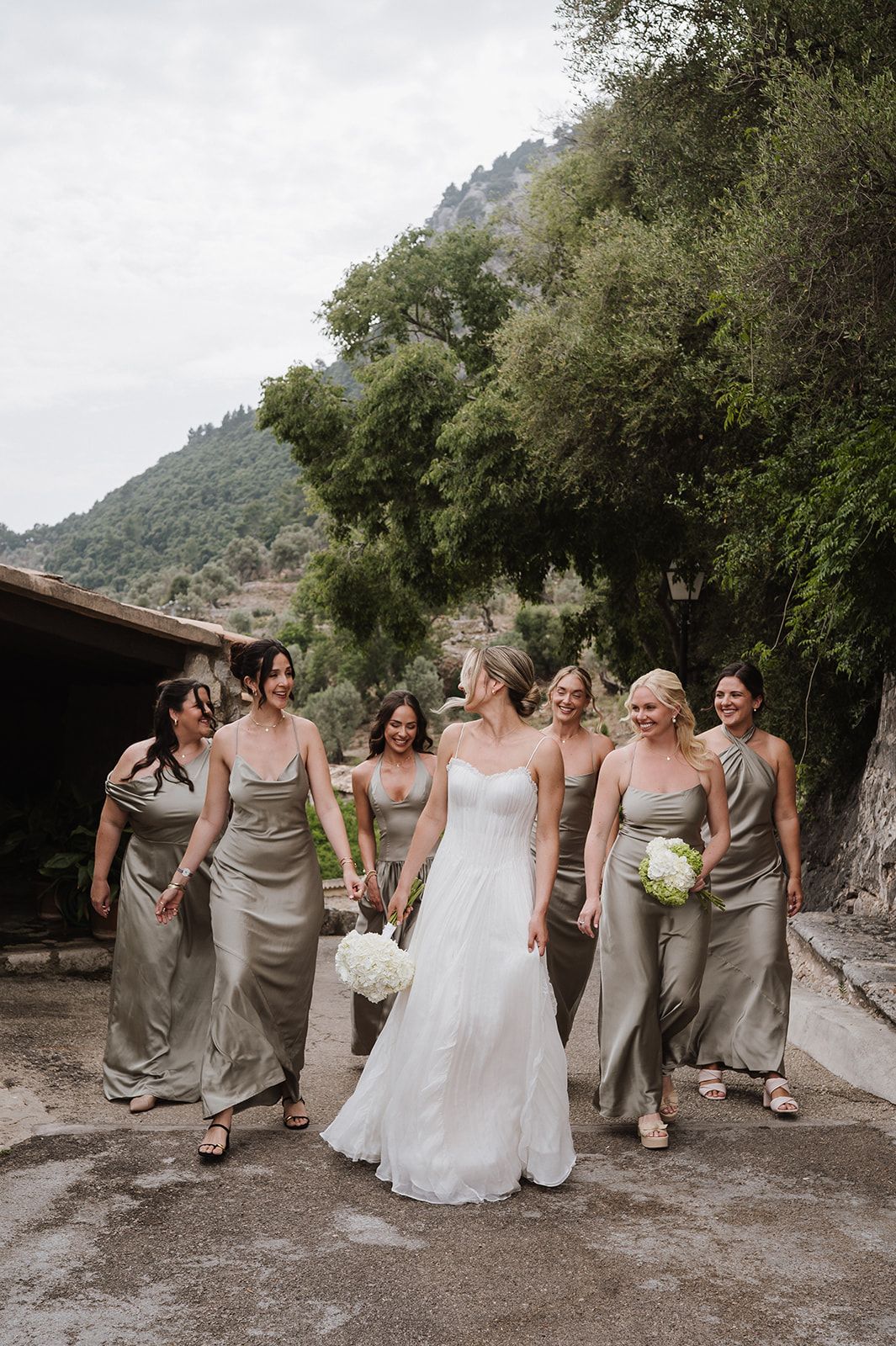 Bride in white gown walks with six bridesmaids in sage green satin dresses against a scenic mountain backdrop.
