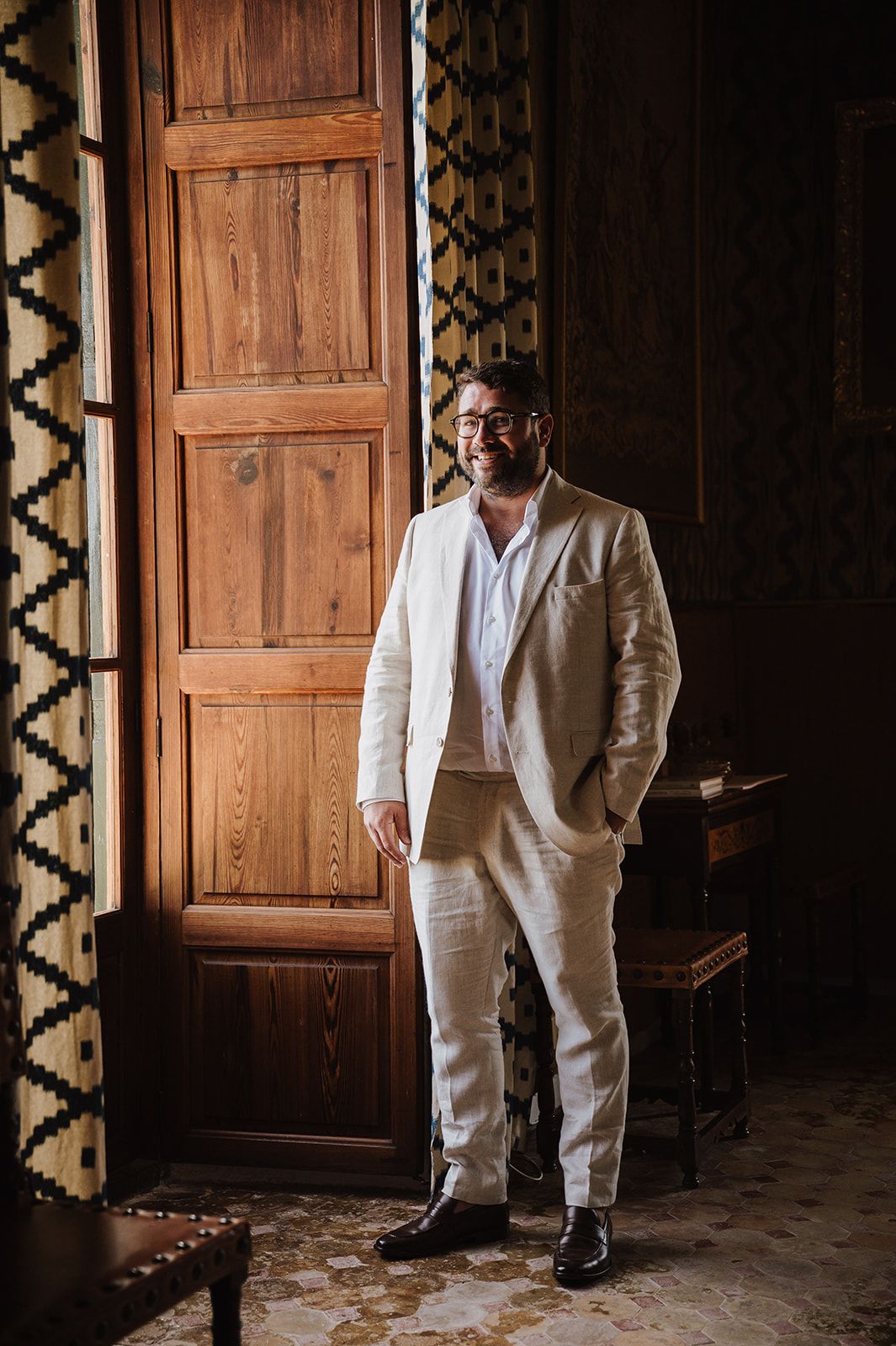 A groom in a beige linen suit smiles while standing by a wooden door in an elegant interior.
