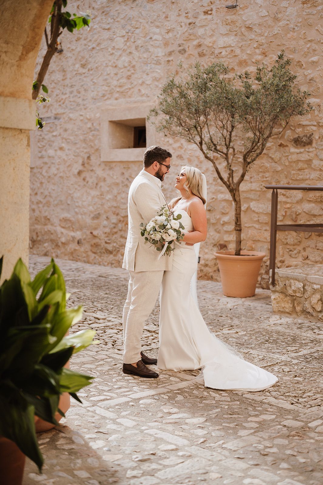 Bride and groom sharing intimate moment in cobblestone courtyard with olive tree backdrop.