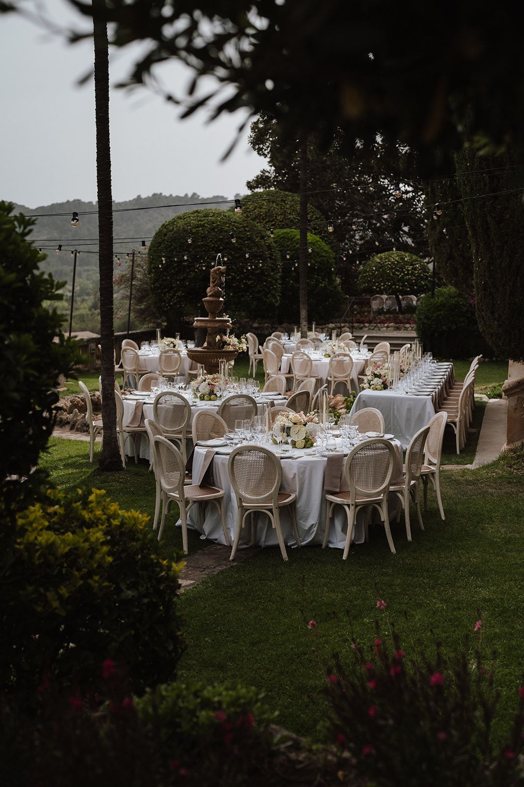 Elegant outdoor wedding reception with round tables, white chairs, and fountain centerpiece in lush garden setting.