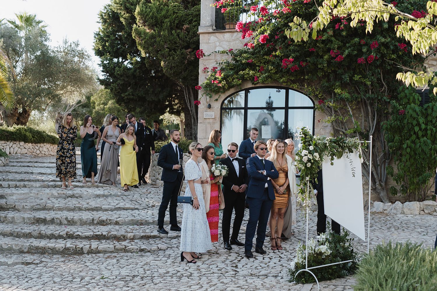 Wedding guests gather on cobblestone steps surrounded by bougainvillea at a scenic outdoor venue.