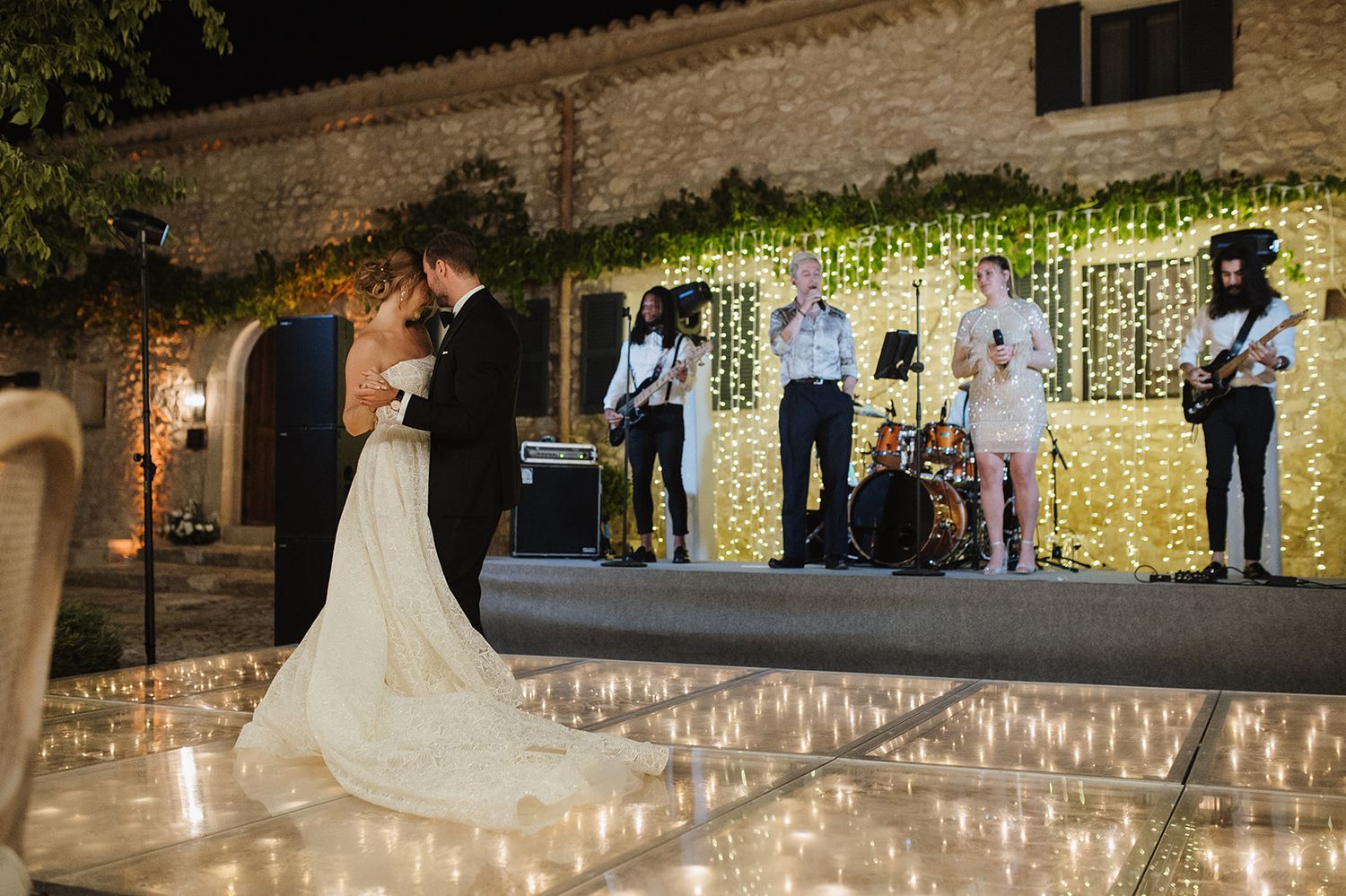 Bride and groom share first dance on mirrored floor while live band performs on a fairy-lit stage.