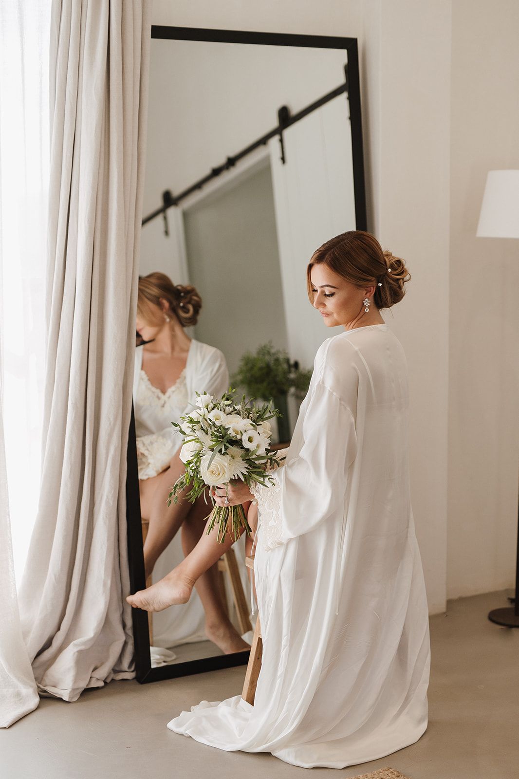 Bride in white robe holds bouquet of white flowers while sitting beside a large floor mirror.