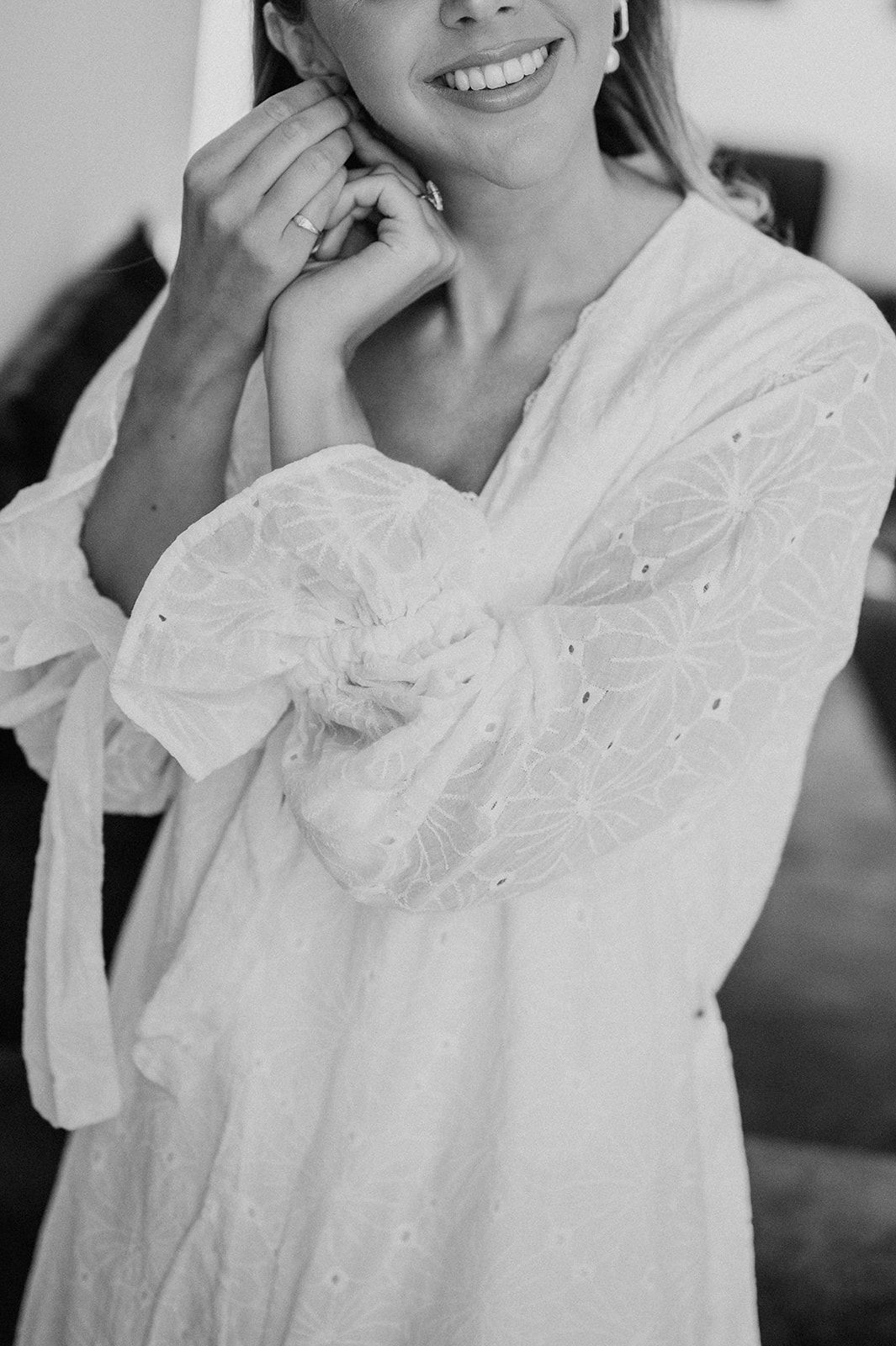 Smiling bride in white broderie anglaise robe adjusting her earring in a black and white photo.