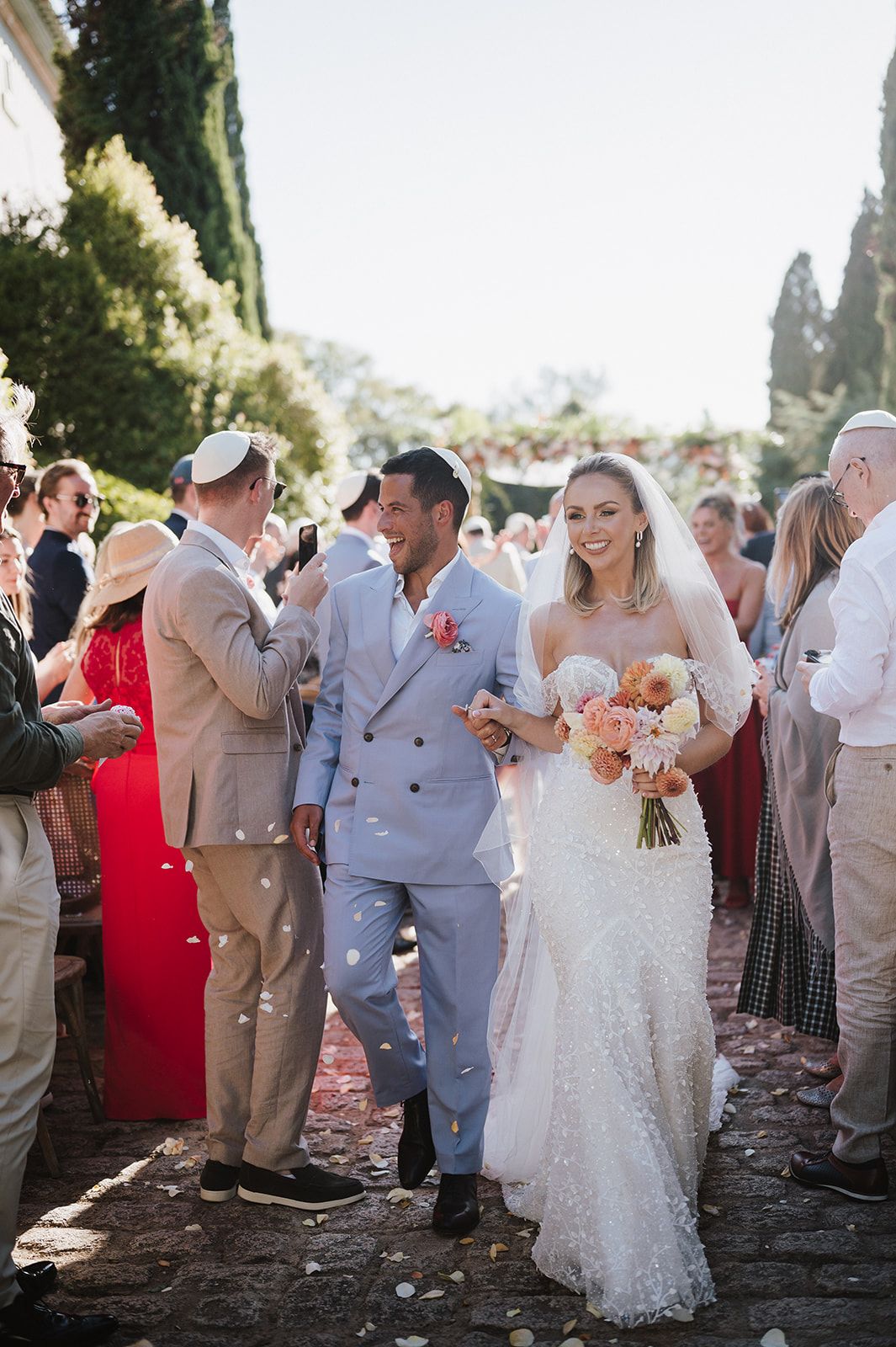 Newlyweds walk down aisle as guests shower them with white confetti during sunny outdoor wedding ceremony.