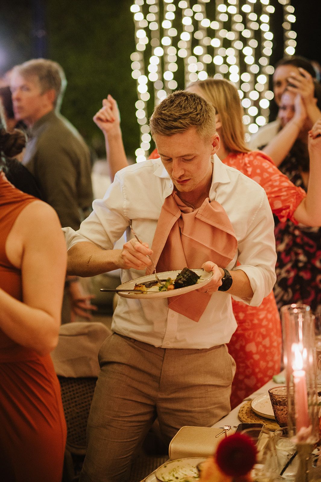 A young man in a white shirt with pink tie focuses on eating at an outdoor wedding reception.