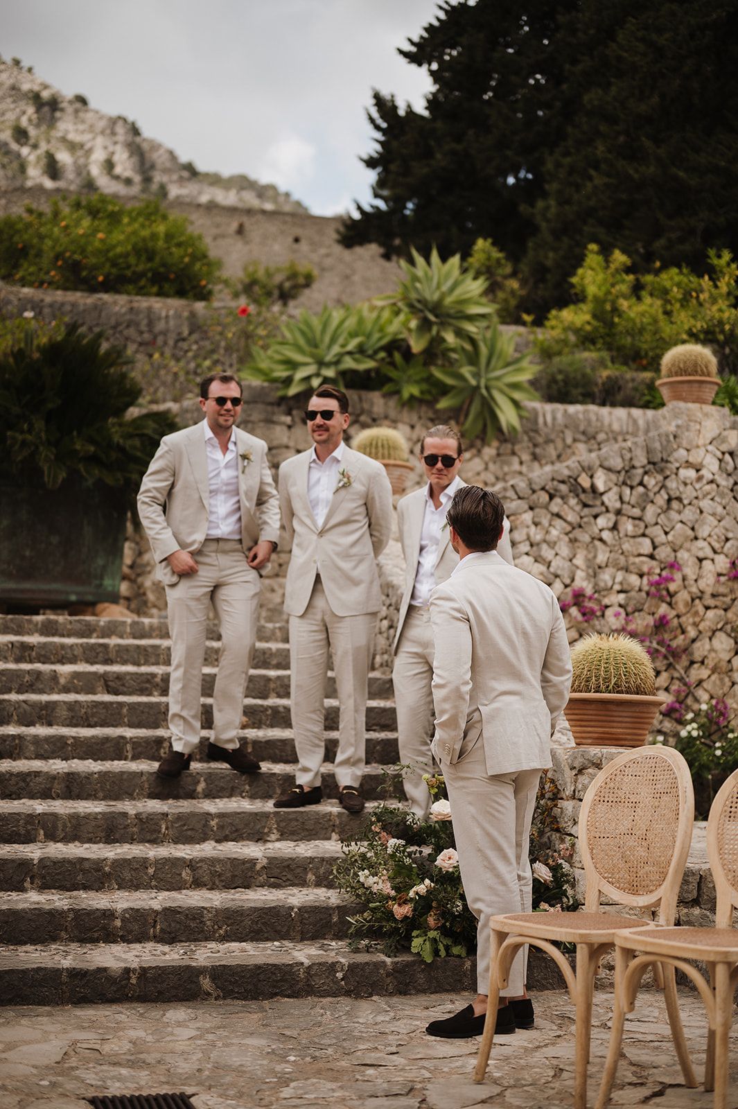 Groomsmen in matching beige linen suits descend stone steps at Mediterranean outdoor wedding venue.