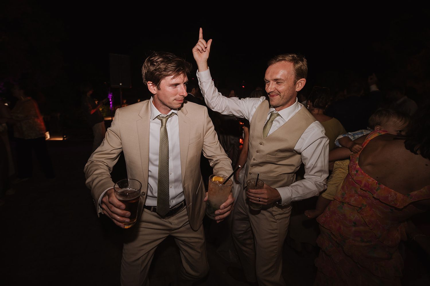 Two men in tan suits dancing and celebrating at an outdoor wedding reception at night.