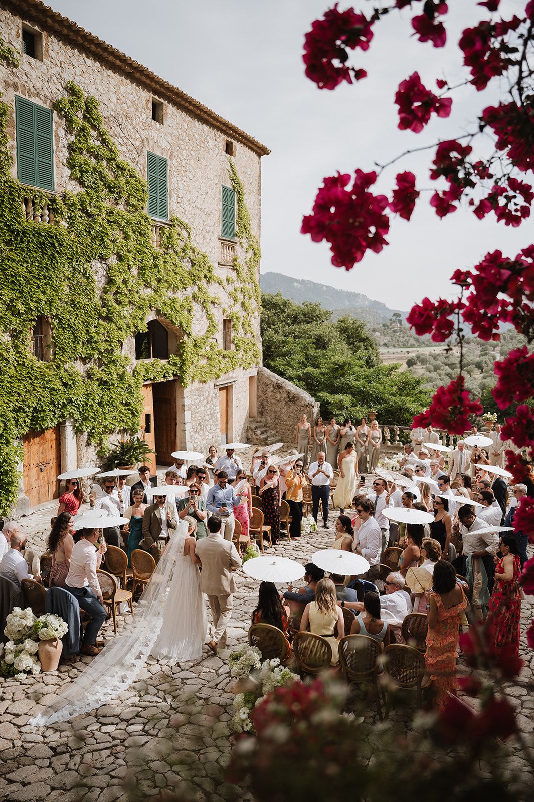 Aerial view of elegant outdoor wedding ceremony at ivy-covered stone villa with guests holding white parasols.