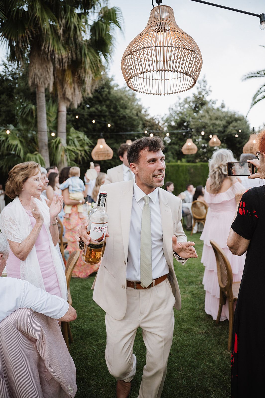 Groom in beige suit holding whiskey bottle at outdoor wedding reception with rattan pendant lights and palm trees.