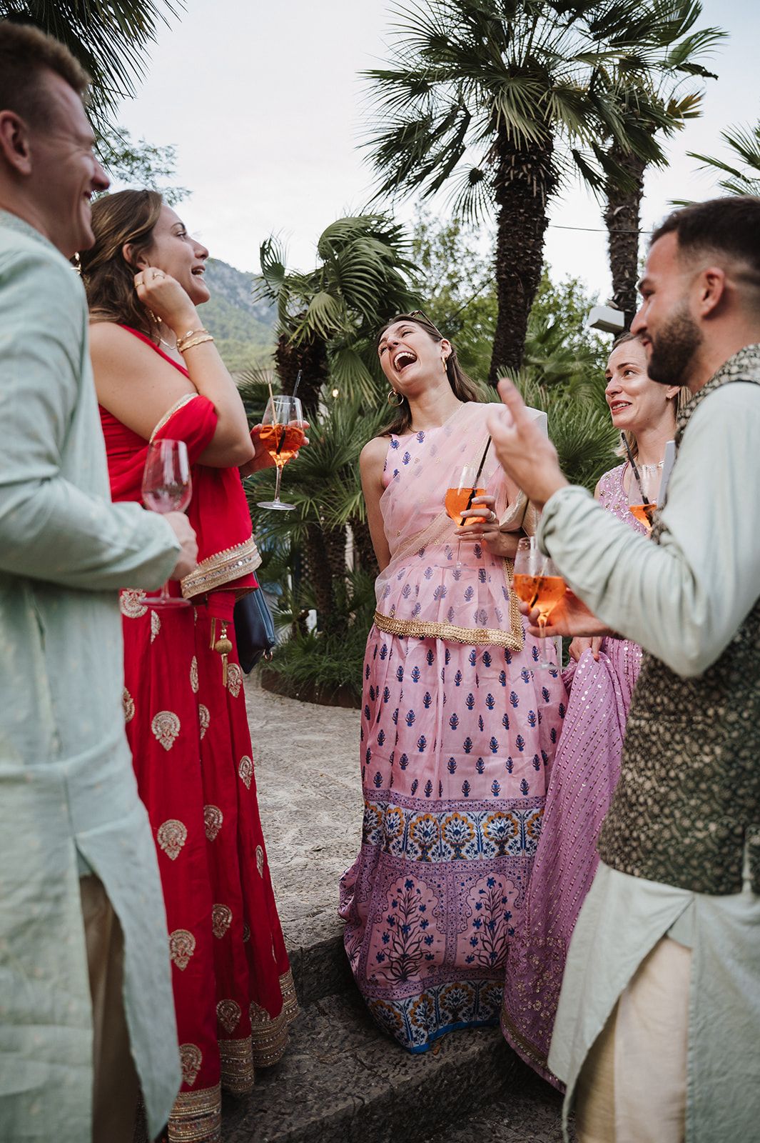 Wedding guests in Indian attire laughing and socializing outdoors with palm trees in background.