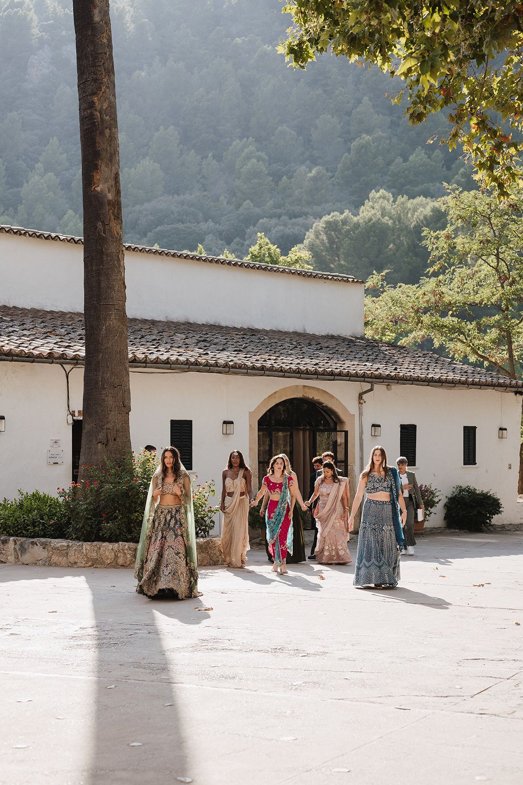 Bridesmaids in colorful Indian lehengas walking across a sunlit courtyard at a Spanish villa wedding.