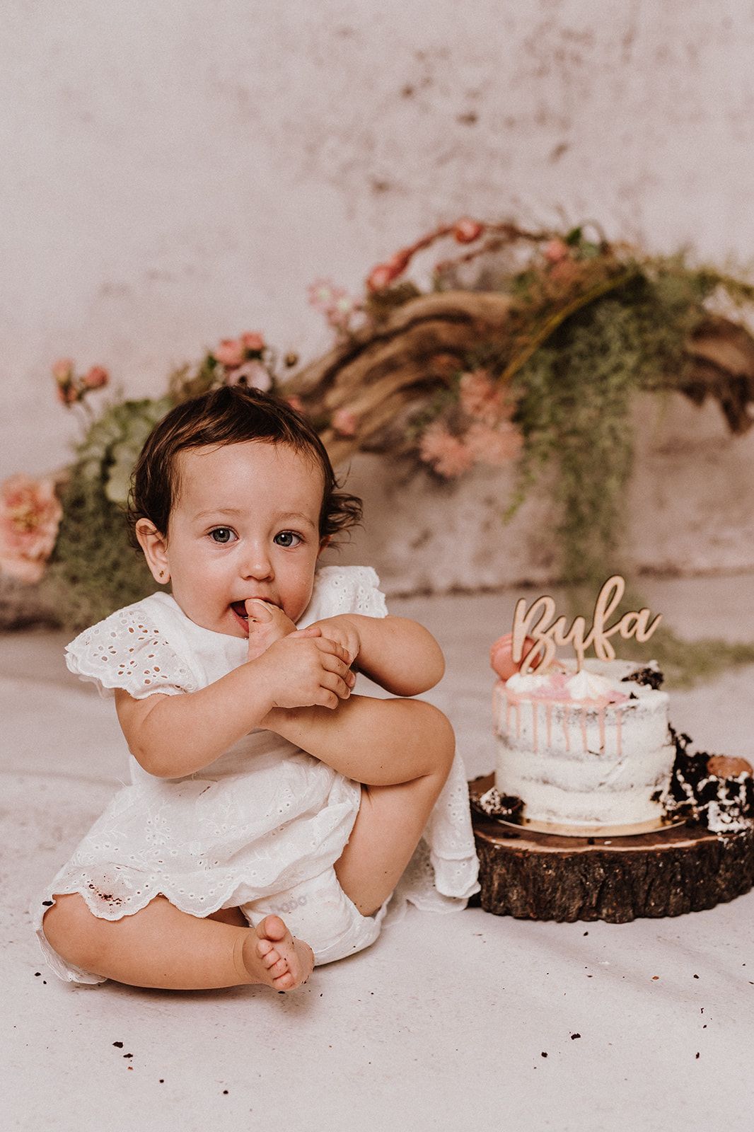 Baby girl in white dress sits beside a small birthday cake with a name topper at a floral first birthday cake smash.