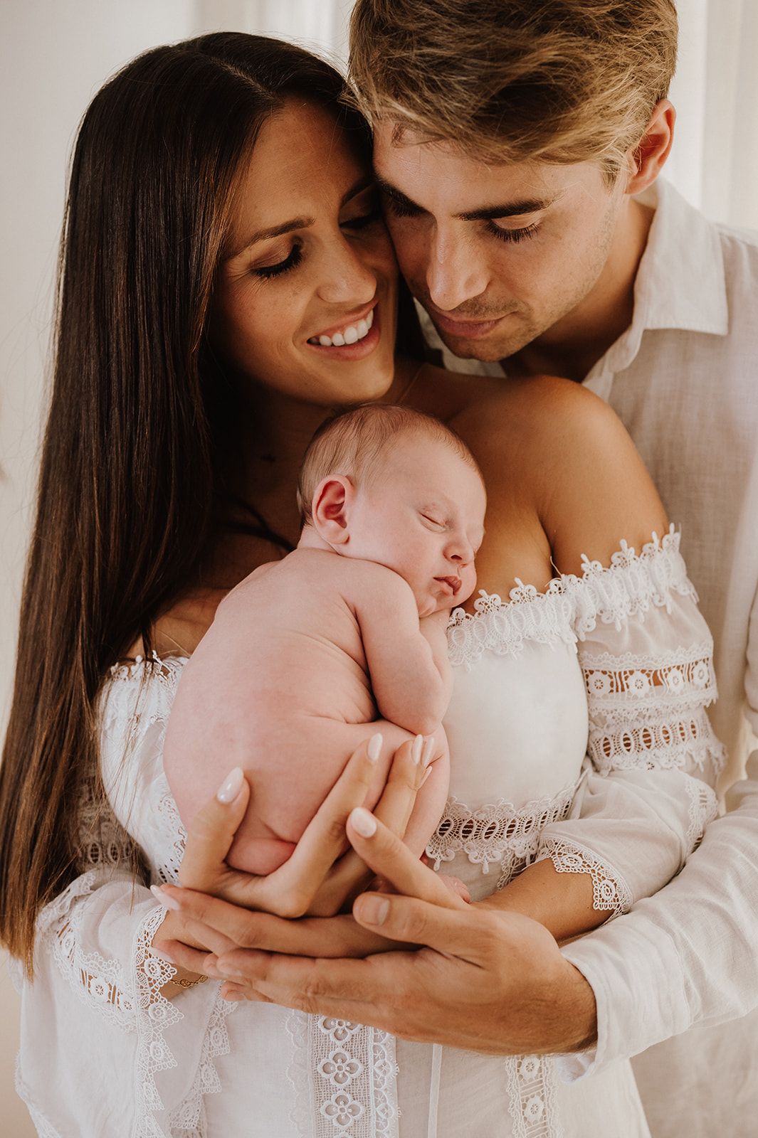 Young couple in white outfits tenderly holding their sleeping newborn baby in a bright, airy setting.
