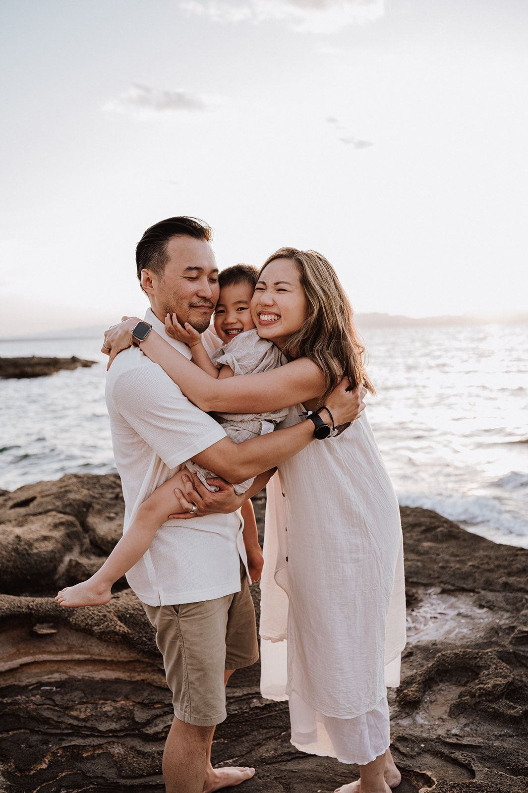 A happy family of three embraces on rocky shoreline at sunset, parents holding young child with ocean in background.