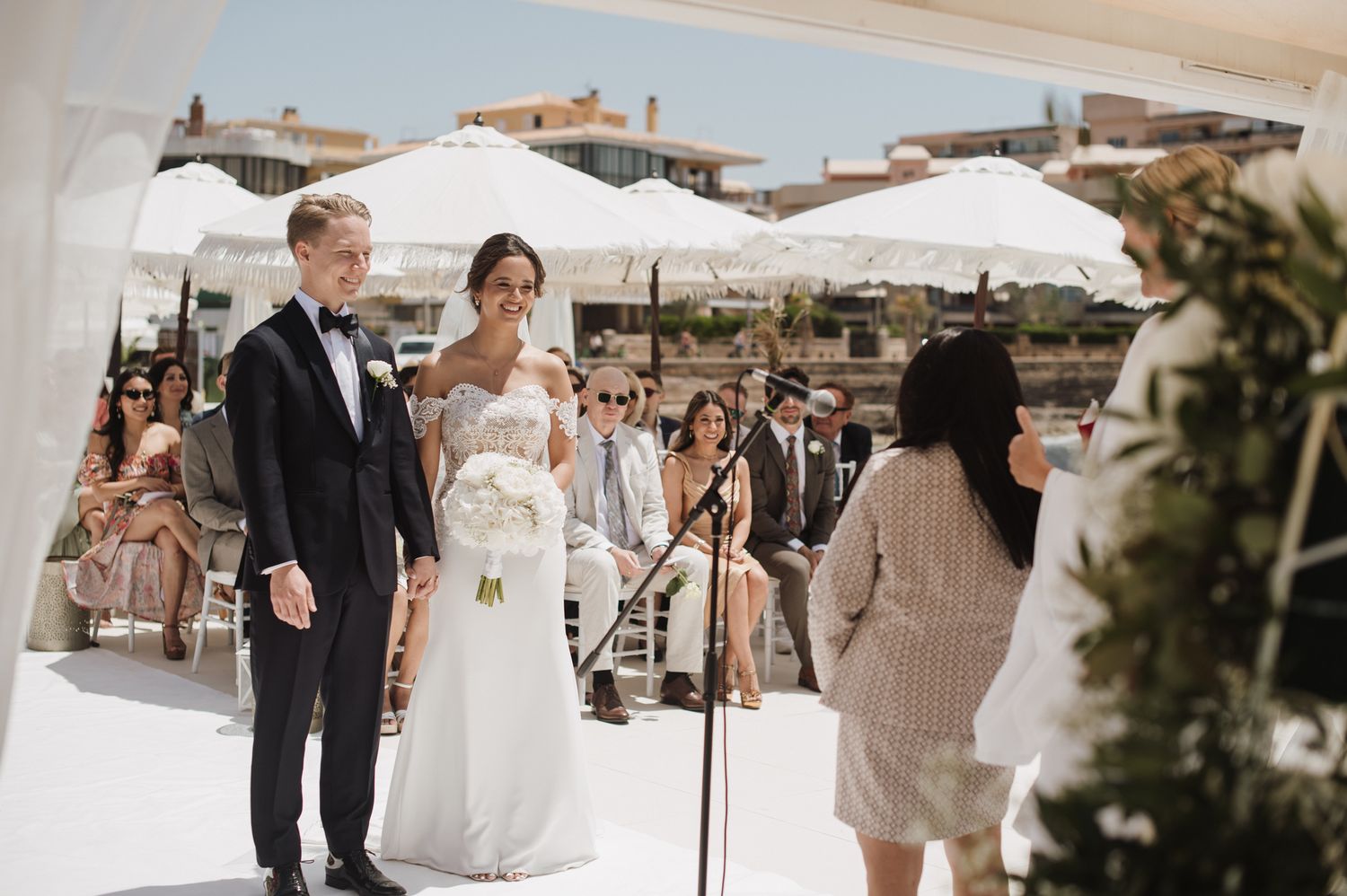 Bride and groom stand at outdoor seaside wedding ceremony altar with guests seated behind.