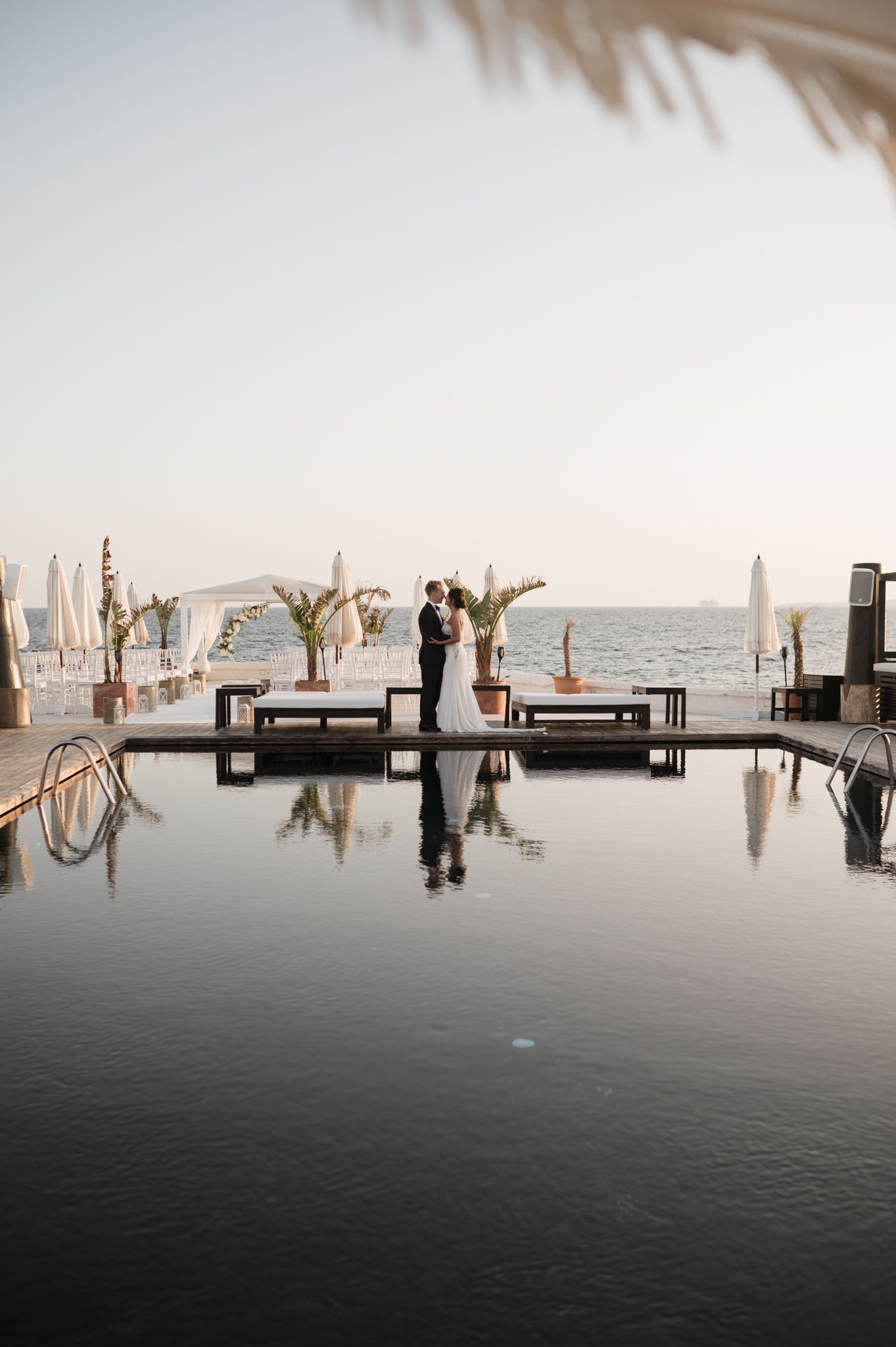 Bride and groom share romantic moment reflected in infinity pool at sunset oceanfront wedding venue.