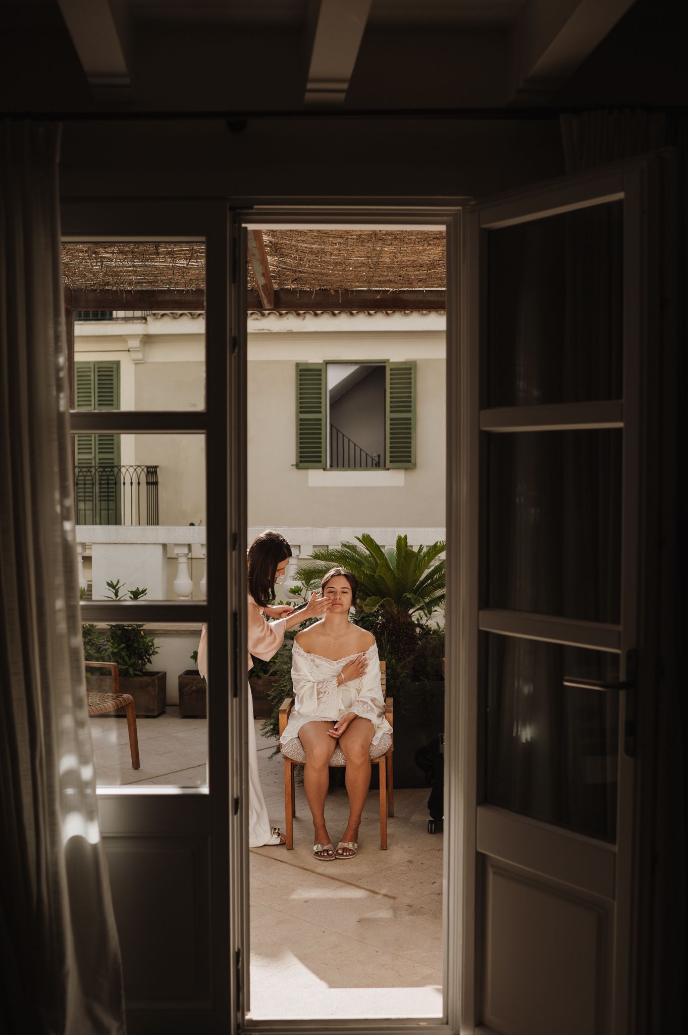 Bride getting makeup done on a sunlit balcony, framed by open French doors with green shutters.