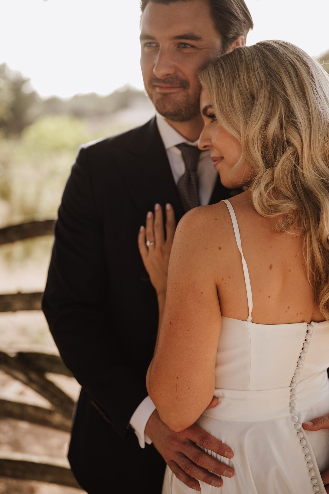 Groom tenderly embracing bride from behind near rustic wooden fence during golden hour wedding portraits.