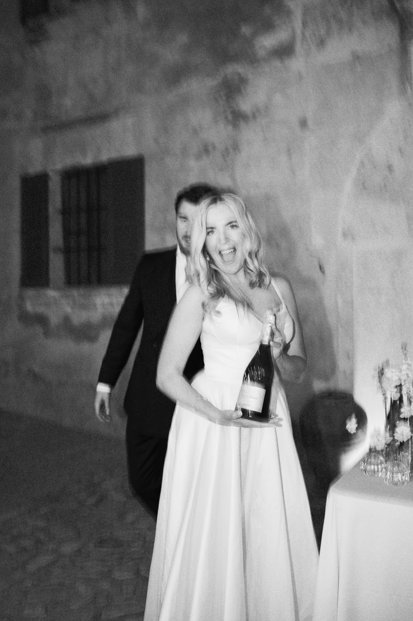 Black and white photo of a laughing bride holding a champagne bottle at her wedding.