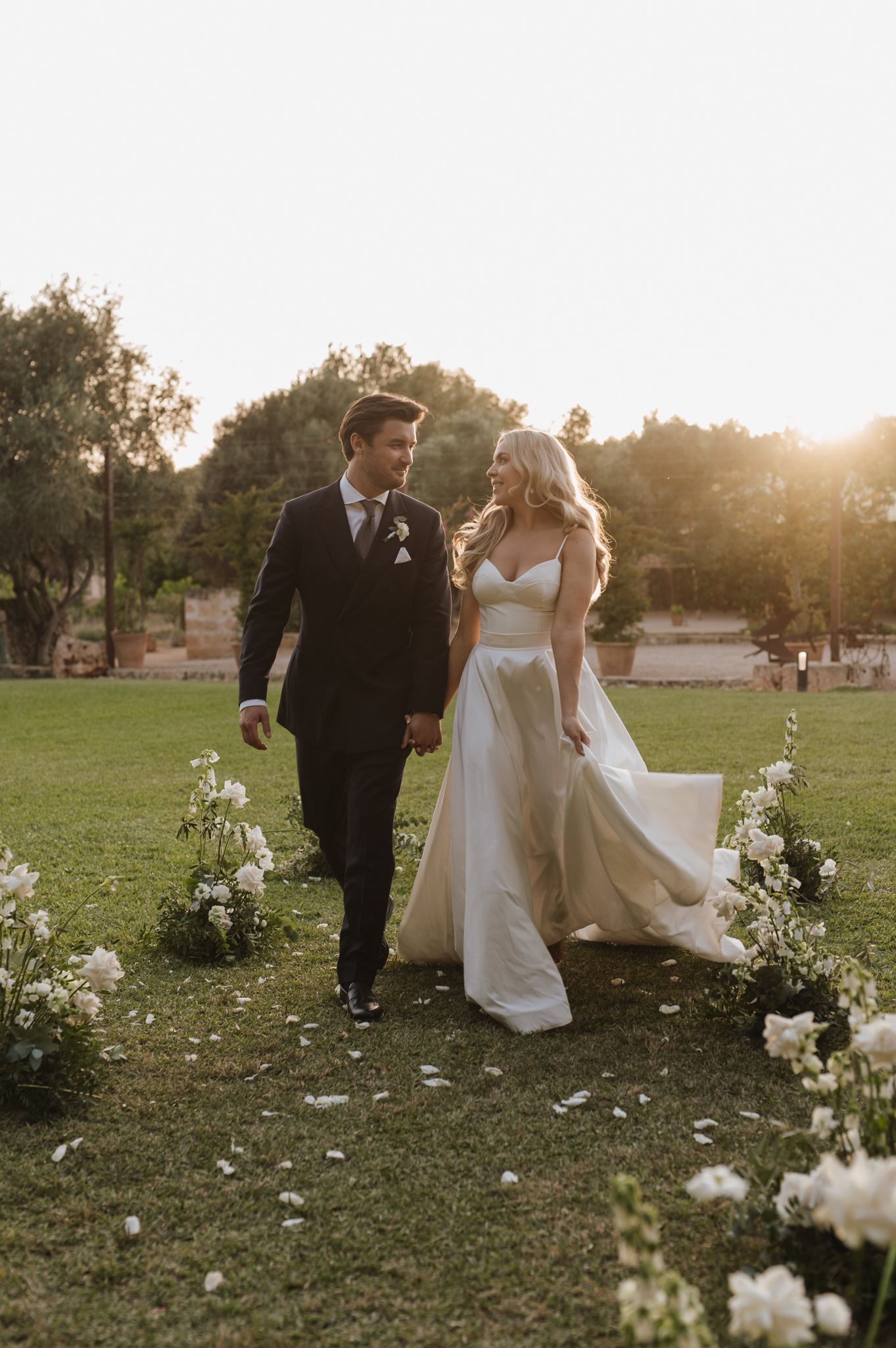 Bride and groom walk hand-in-hand down floral aisle at golden hour outdoor wedding ceremony.