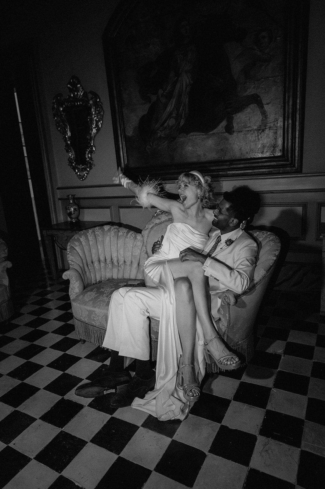 Black and white photo of newlyweds celebrating playfully on a vintage sofa in elegant interior.