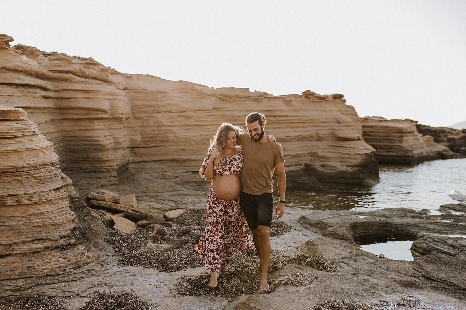 Pregnant couple walking on rocky coastal cliffs, woman in floral dress, man in tan shirt, ocean backdrop.