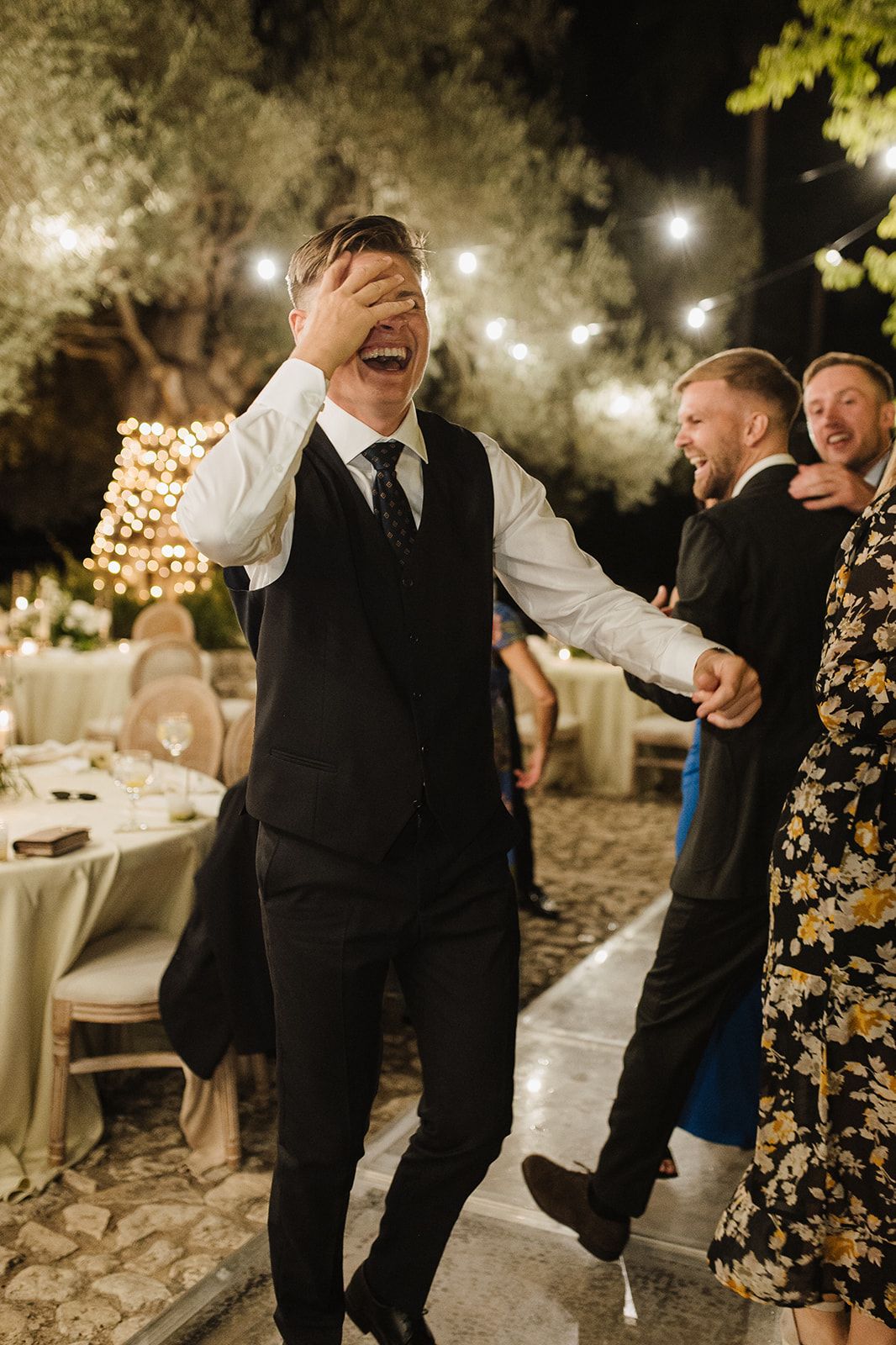 Laughing groomsman covers his face while dancing at a festive outdoor wedding reception under string lights.