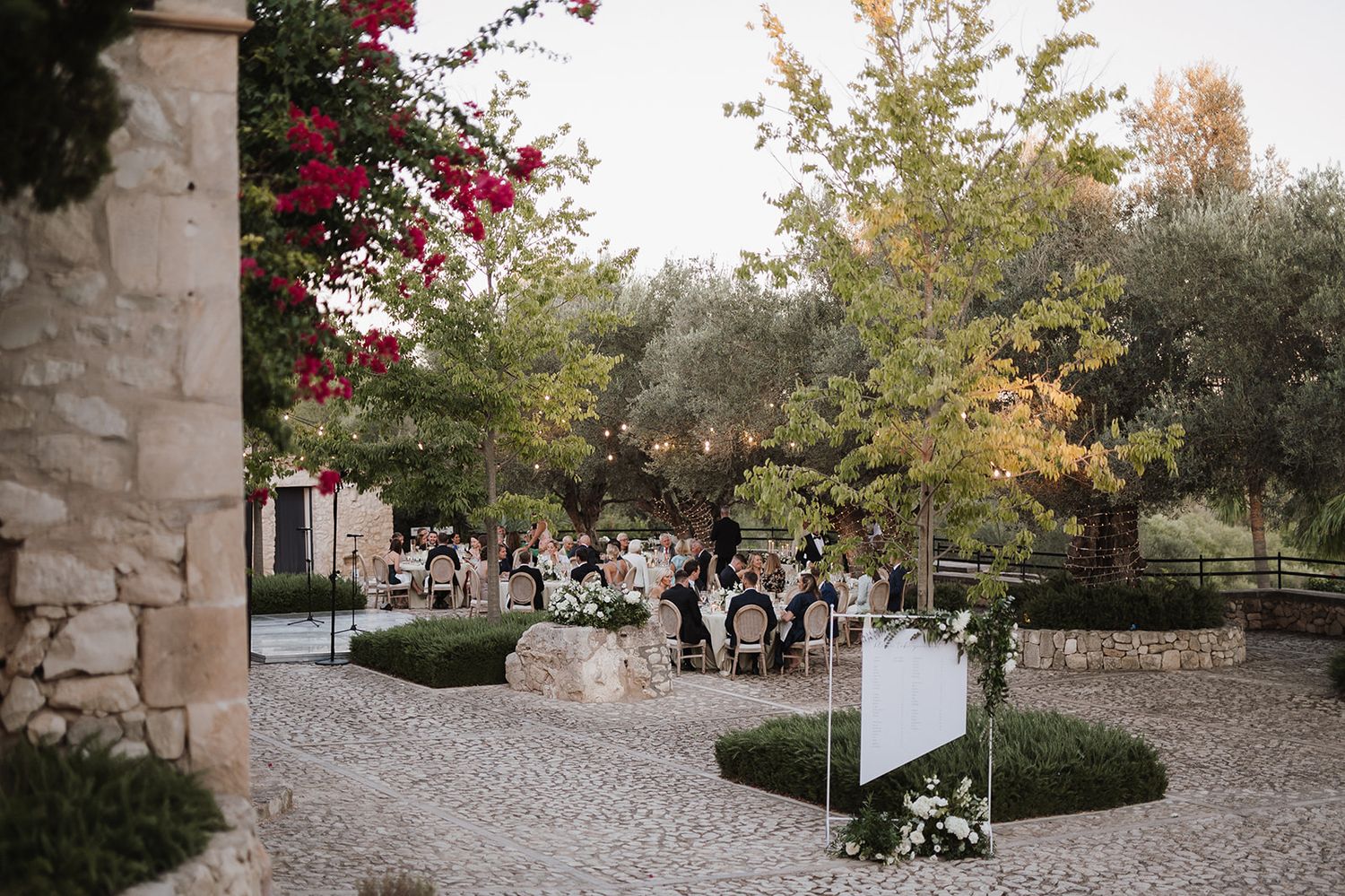 Elegant outdoor wedding reception in a stone courtyard with string lights, lush trees, and floral arrangements at dusk.