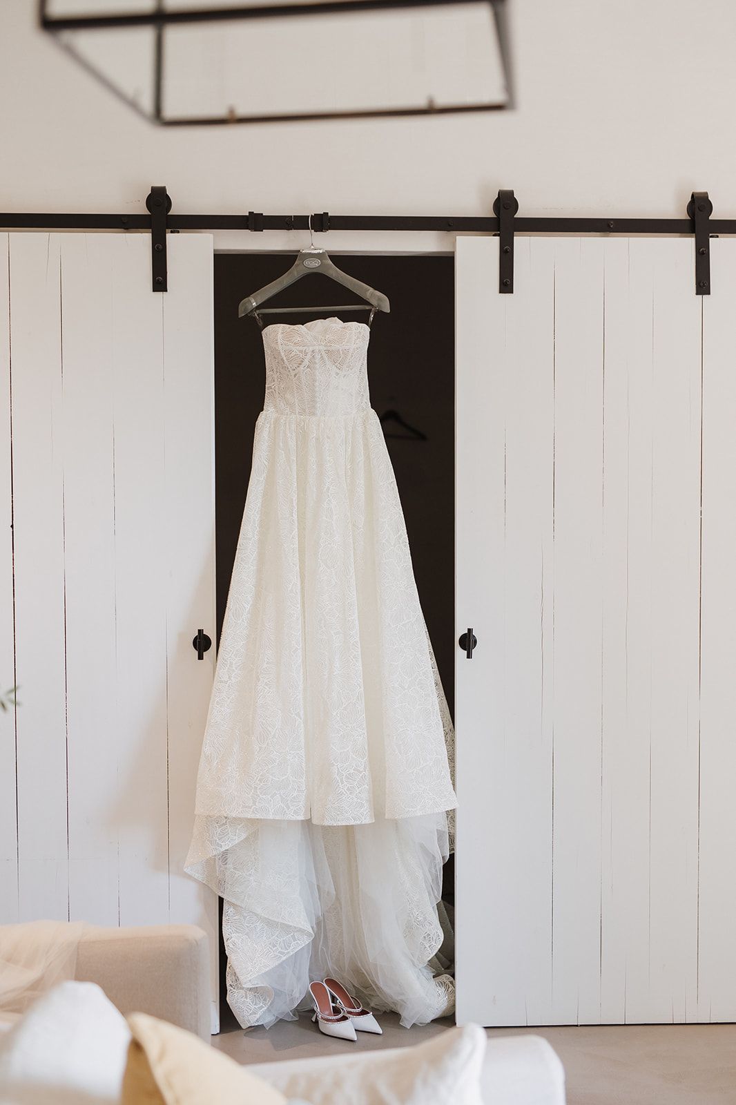 Elegant strapless white wedding dress hanging on rustic barn door with bridal heels below.
