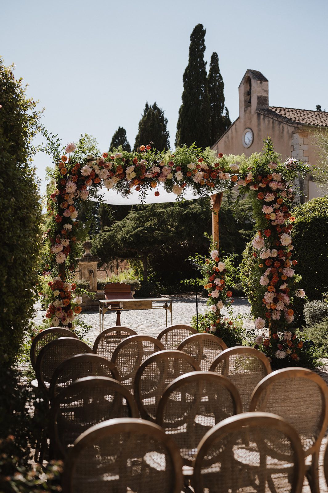 Outdoor wedding ceremony arch adorned with pink and coral flowers, surrounded by cypress trees.