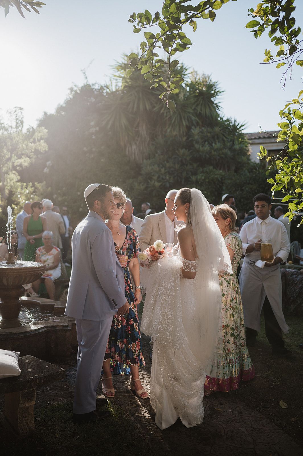 Bride in lace veil and groom in light grey suit greeting guests by a stone fountain in sunlit garden.