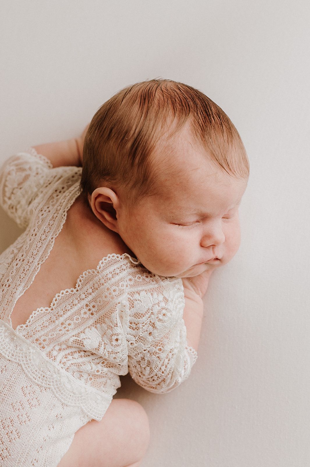 Sleeping newborn baby in white lace romper lying peacefully on a soft white background.